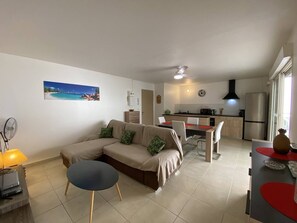 Ein modernes Wohnzimmer mit einem beigefarbenen Sofa, einem kleinen runden Tisch und einem Ventilator. Im Hintergrund ist eine Küche mit Kühlschrank, Herd und Essbereich zu sehen.