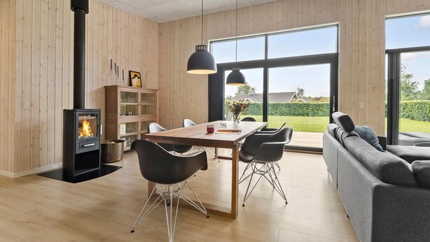 Een moderne woonkamer met een houtkachel, een eettafel met stoelen, een bank en grote ramen met uitzicht op het landschap.