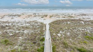 Ein Strand mit einer Holzpromenade, die zum Wasser führt, umgeben von Dünen und vereinzelter Vegetation.