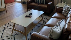 A living room with a brown leather sofa, a white coffee table, and a ceiling fan. There are windows with curtains, a door, and a small table with a plant.
