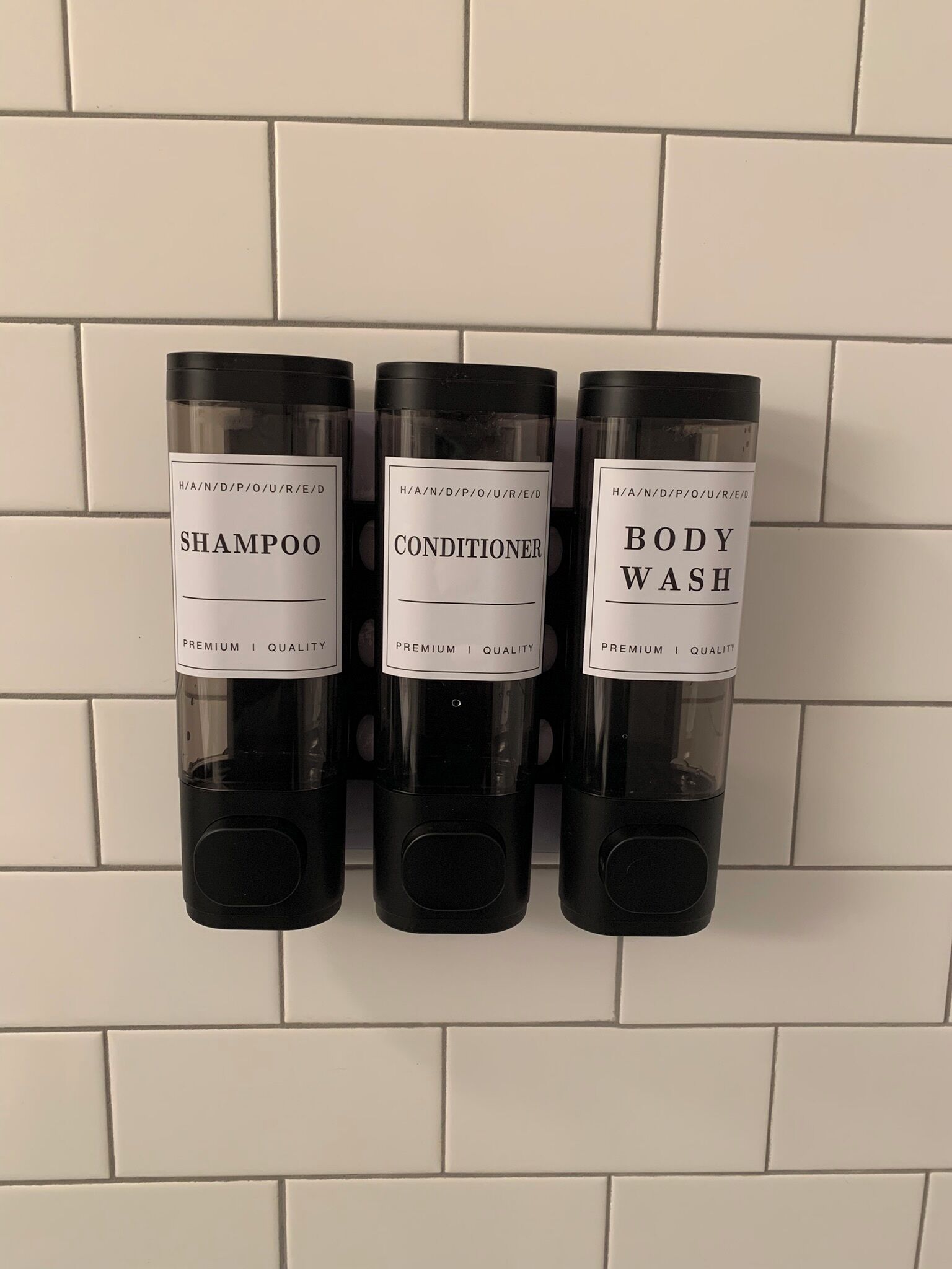 Three black dispensers labeled 'SHAMPOO', 'CONDITIONER', and 'BODY WASH' mounted on a white tiled wall.