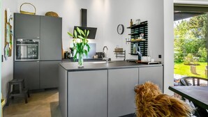 A modern kitchen with grey cabinets, a built-in oven, a large island with a vase of green plants, and a view of a garden through a glass door. - "Melina" - 900m to the fjord by Interhome (Farsø)
