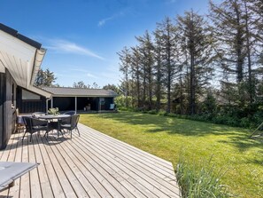 Une terrasse en bois avec des meubles d’extérieur, un patio couvert et un paysage boisé en arrière-plan.