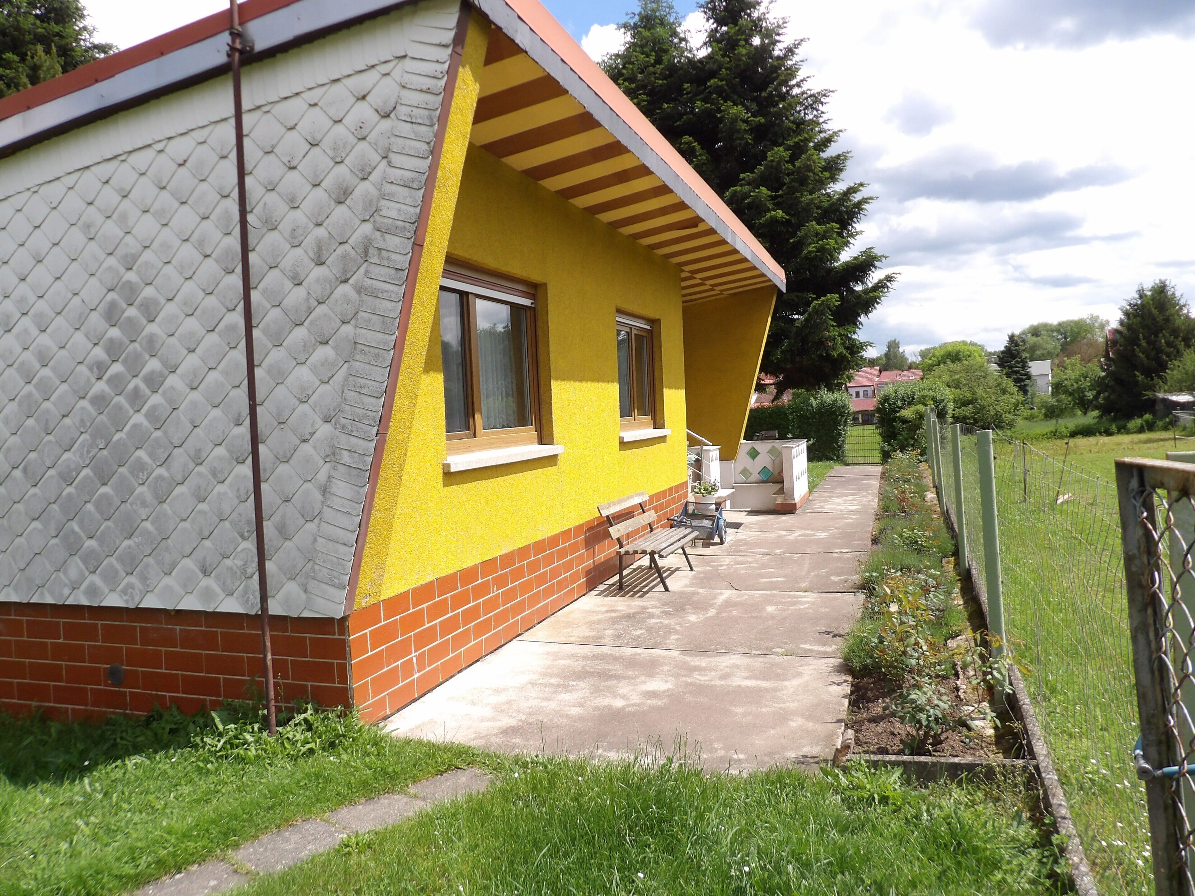 Ein eingeschossiges Haus mit gelber Fassade, einem Ziegelmauerwerk und einer kleinen Veranda mit einer Bank.