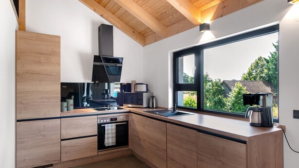 A modern kitchen with wooden cabinetry, a built-in oven, a microwave, and a large window with a view of greenery. - Holiday Apartment 'Ferienoase Im Land Der 1000 Seen' with Private Terrace, Shared Garden & Wi-Fi (Karstädt)