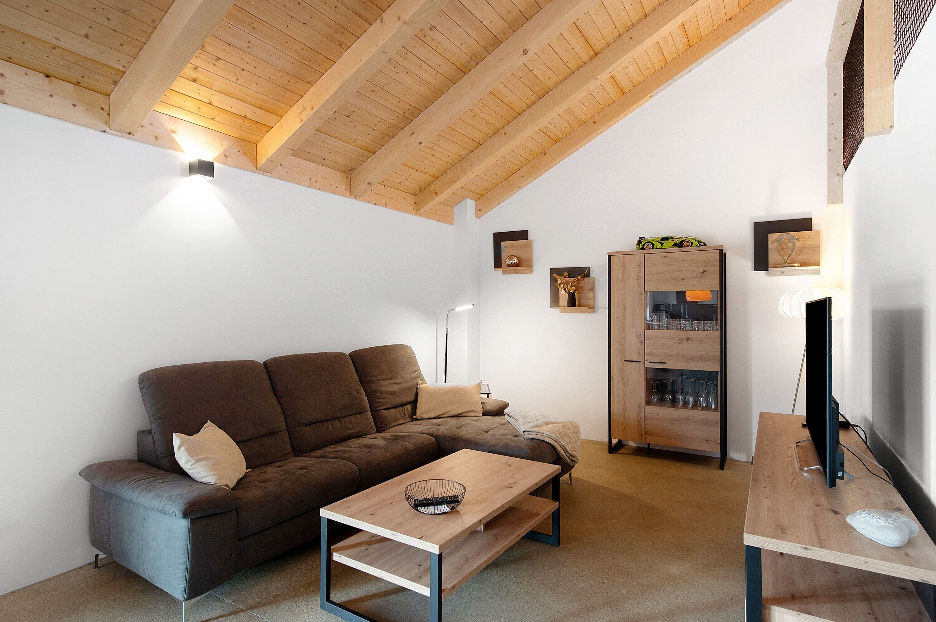 Un salon moderne avec un plafond en bois, un canapé marron, une table basse en bois, un meuble pour la télévision et un buffet contenant de la verrerie.