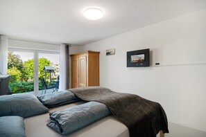A bedroom with a bed, a wardrobe, a desk, and a window with a view of greenery.