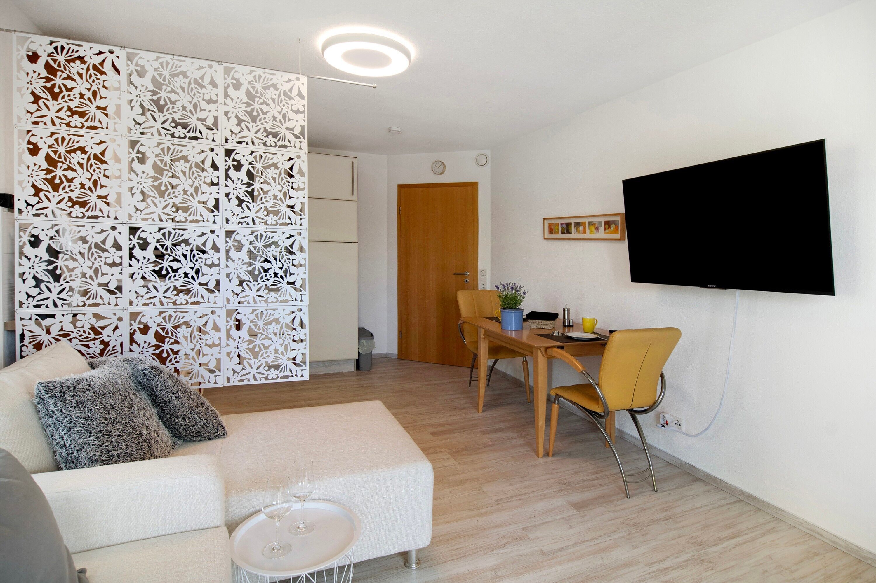 Un soggiorno con un divano, un tavolo da pranzo con sedie, una TV a parete e un pannello decorativo bianco.