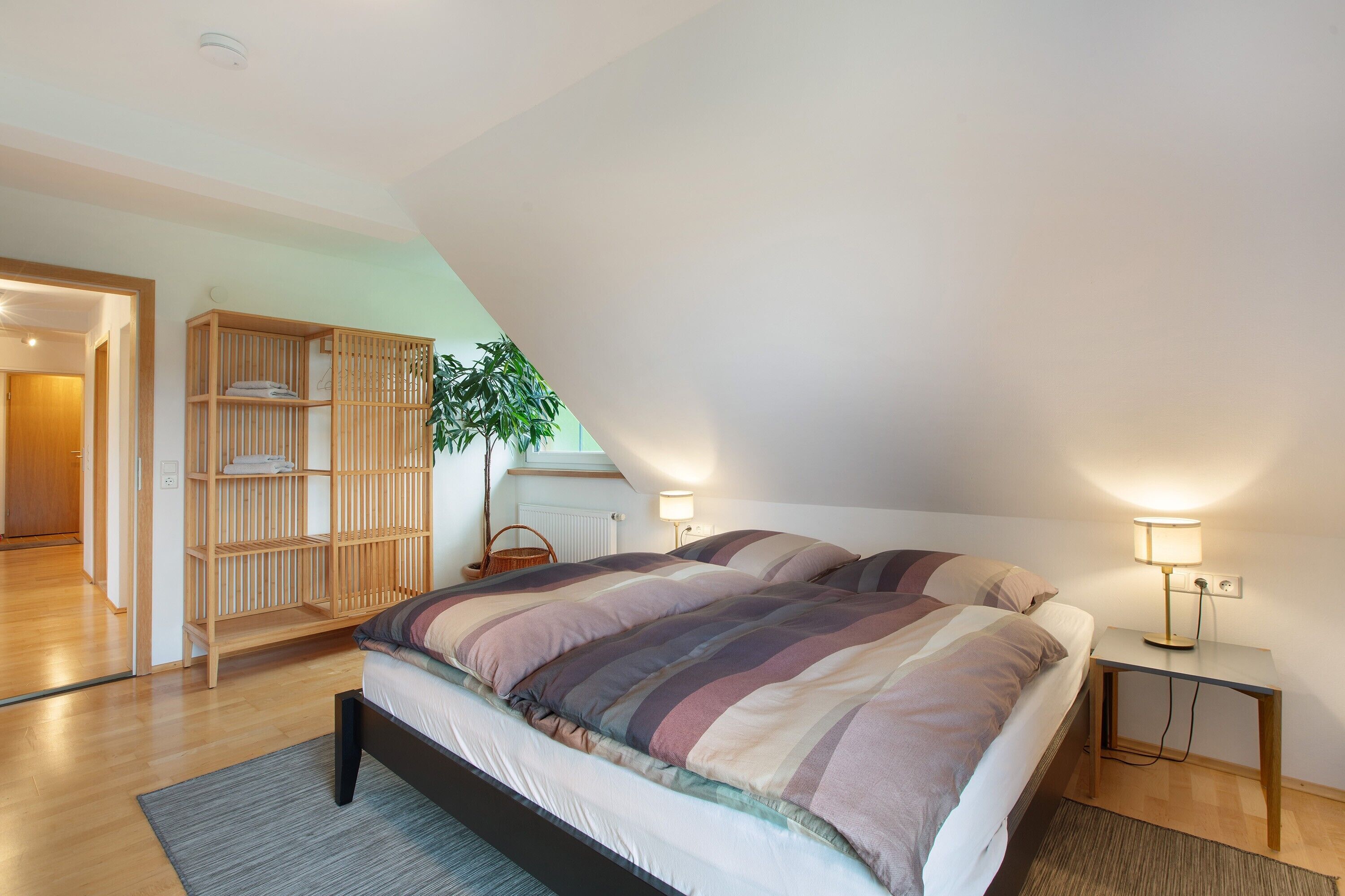 A bedroom with a bed, bedside lamps, a wooden wardrobe, and a plant.