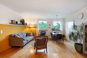 A living room with a sofa, dining table, chairs, a wicker chair, a lamp, a clock, and a potted plant.