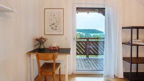 Ein Zimmer mit einem Holztisch, einem Stuhl und einer gerahmten botanischen Illustration. Durch eine Tür ist Blick auf eine hölzerne Terrasse im Freien.