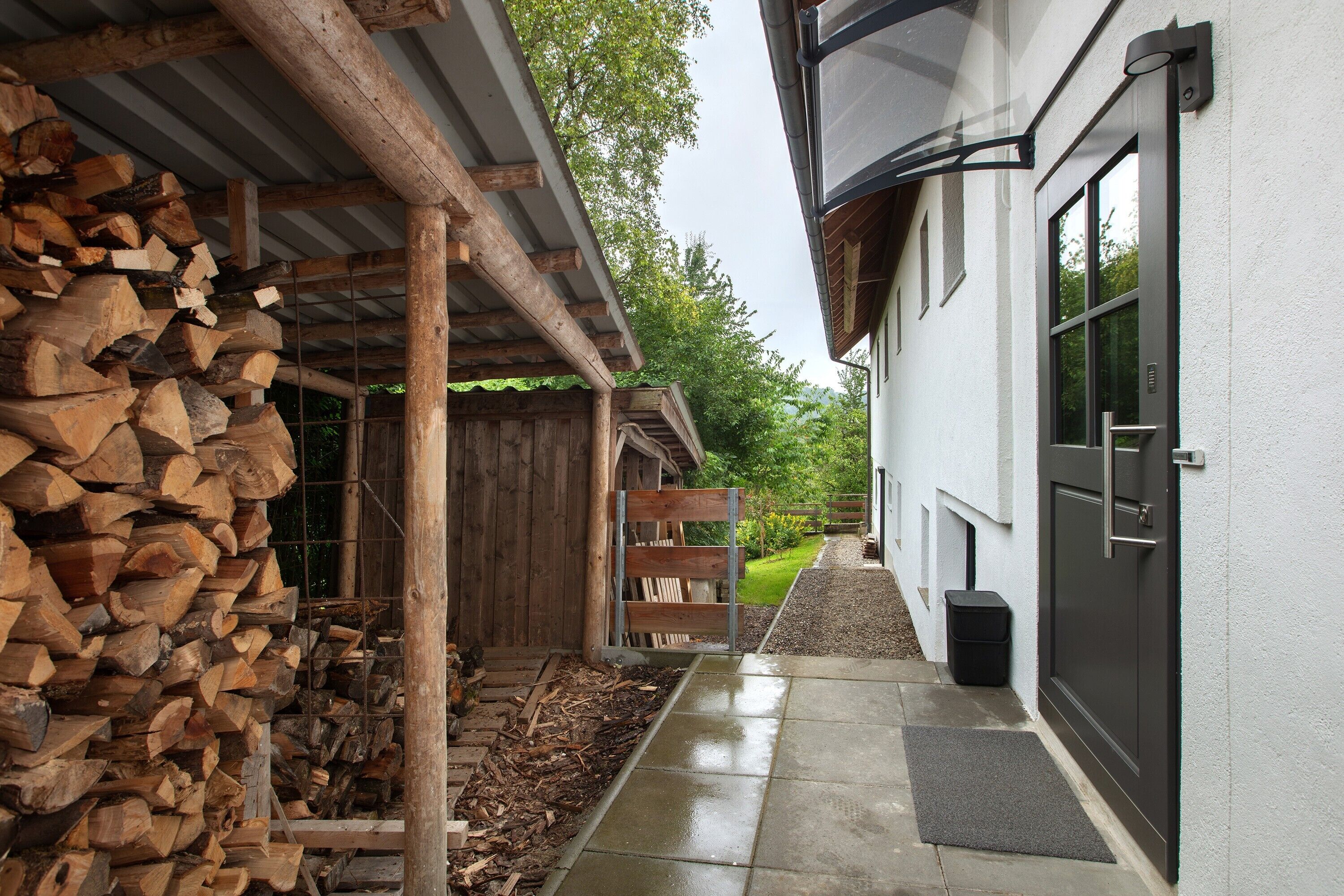 A wooden structure with a pile of firewood, a metal gate, and a concrete pathway leading to a building with a black door and a window.