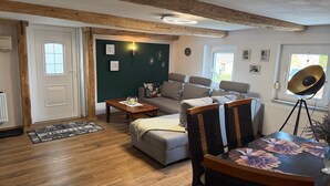 En hyggelig stue med sofa, sofabord, spisebord med en vase, og et stort vindue med udsigt.