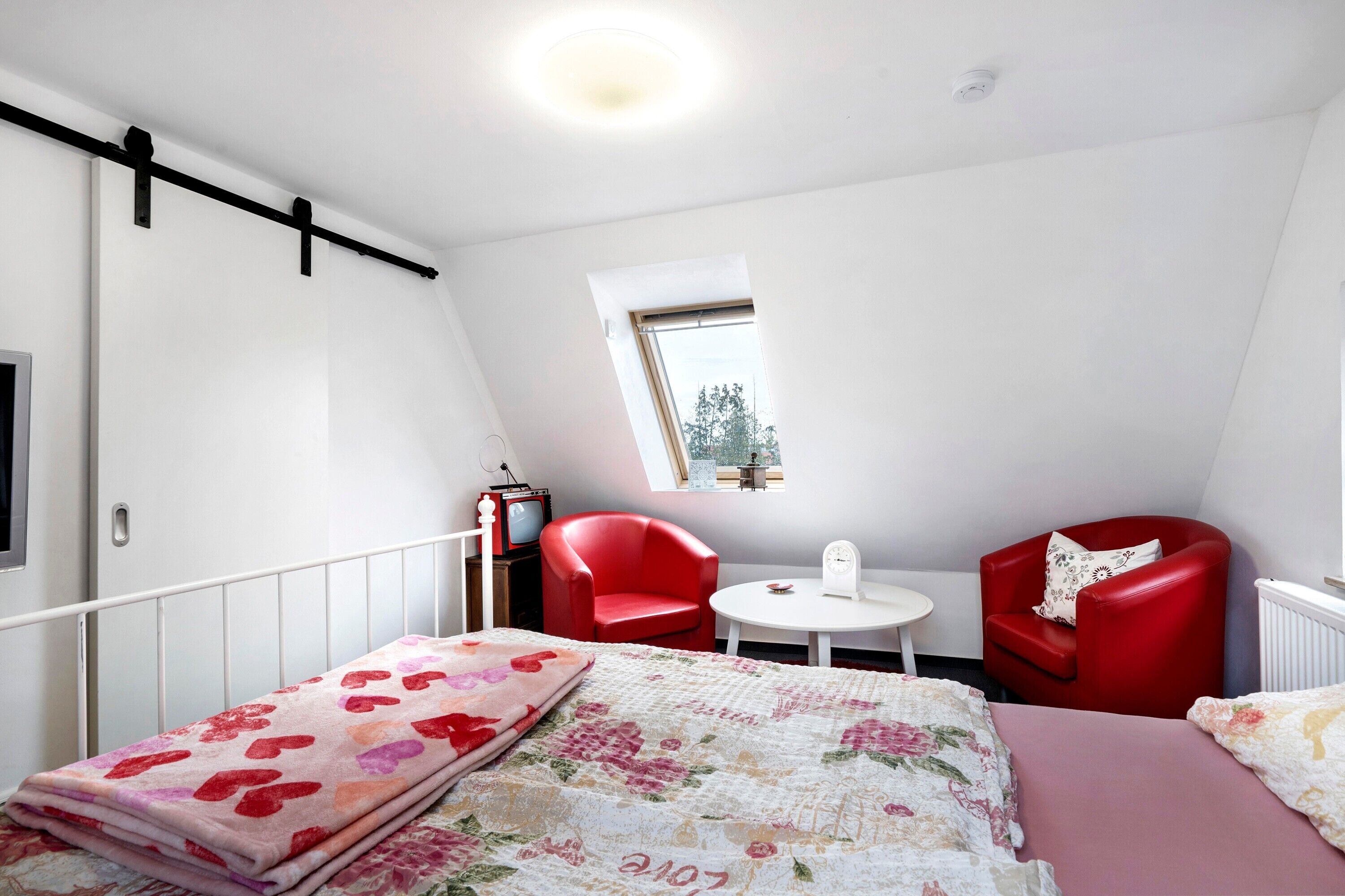 A bedroom with a bed, two red chairs, a small table, a window, and a radiator.