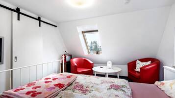 A bedroom with a bed, two red chairs, a small table, a window, and a radiator.