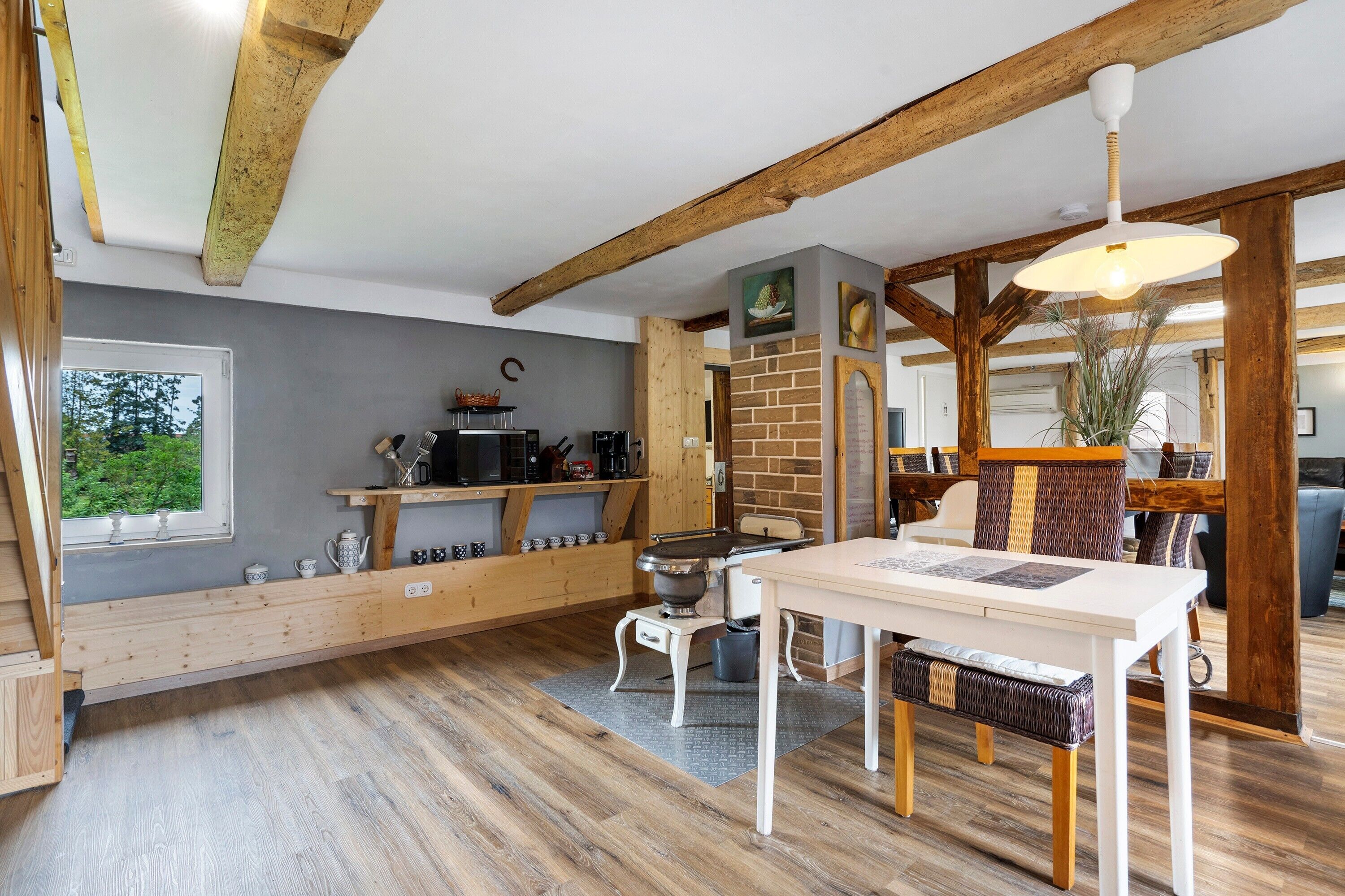 A rustic kitchen with wooden beams, a dining area with a table and chairs, and a view of greenery outside.