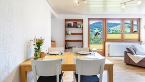 A dining area with a wooden table and chairs, a bookshelf, and a view of a green landscape through large windows. - Holiday Apartment 'Käppeleblick' with Mountain View, Private Terrace and Wi-Fi (Bernau im Schwarzwald)