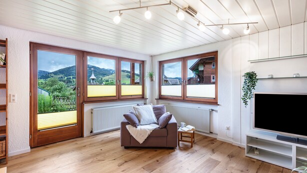 A modern living room with a sofa, TV, and large windows overlooking a scenic landscape. - Holiday Apartment 'Käppeleblick' with Mountain View, Private Terrace and Wi-Fi (Bernau im Schwarzwald)