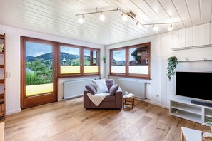 A modern living room with a sofa, TV, and large windows overlooking a scenic landscape. - Holiday Apartment 'Käppeleblick' with Mountain View, Private Terrace and Wi-Fi (Bernau im Schwarzwald)