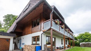 A two-story house with a wooden exterior, a balcony with flower planters, a visible garage, and a well-maintained lawn. - Holiday Apartment 'Käppeleblick' with Mountain View, Private Terrace and Wi-Fi (Bernau im Schwarzwald)