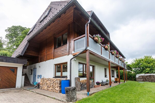 A two-story house with a wooden exterior, a balcony with flower planters, a visible garage, and a well-maintained lawn. - Holiday Apartment 'Käppeleblick' with Mountain View, Private Terrace and Wi-Fi (Bernau im Schwarzwald)