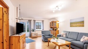 A living room with a TV, wooden furniture, a sofa, and a painting on the wall. - Apartment 'Fereinhof Paulsen - Lachmöwe' with Private Terrace, Shared Garden and Wi-Fi (Pellworm)