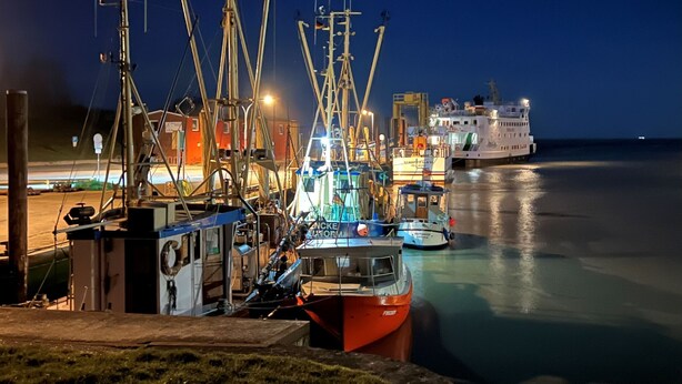A harbor at night with boats docked, a large ship in the background, and a building with visible windows. - Apartment 'Fereinhof Paulsen - Lachmöwe' with Private Terrace, Shared Garden and Wi-Fi (Pellworm)