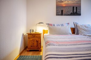 Une chambre à coucher avec un lit, des oreillers, une table de chevet avec une lampe, un plancher en bois et un tableau accroché au mur représentant un coucher de soleil sur l’eau.