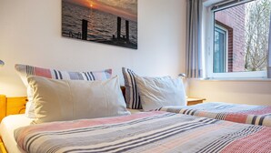 A hotel room with two beds, a painting of a sunset over water, and a window with a view of trees. - Apartment 'Fereinhof Paulsen - Lachmöwe' with Private Terrace, Shared Garden and Wi-Fi (Pellworm)