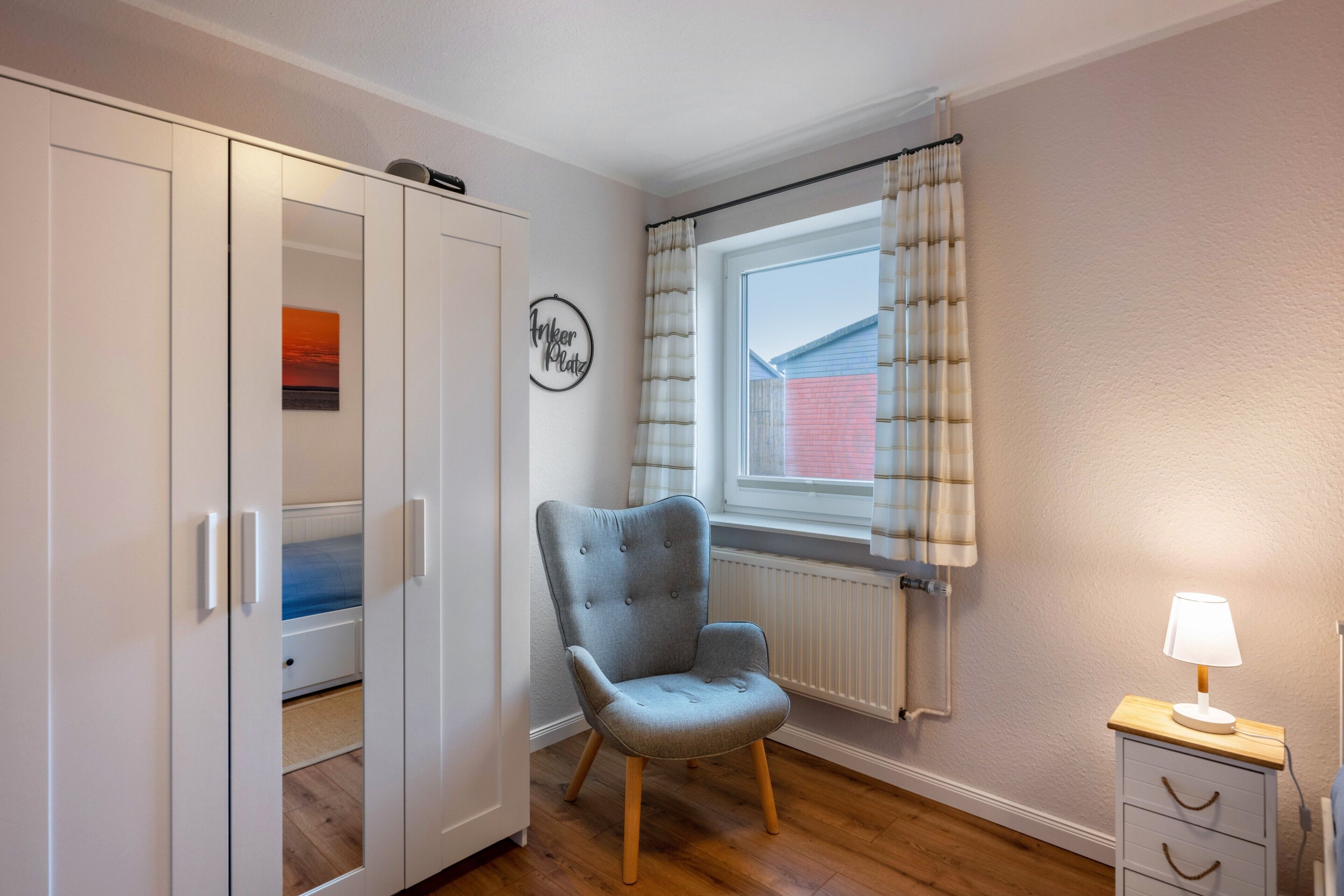 Une chambre avec une armoire, une chaise, une fenêtre avec des rideaux et une table de chevet avec une lampe.