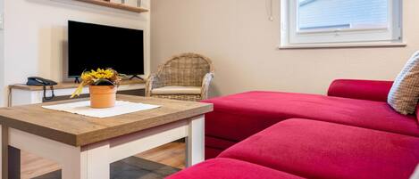 A living room with a flat-screen TV, a wicker chair, a red sofa, a wooden table, and a window with a view of a building.
