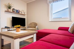 Ein Wohnzimmer mit einem Flachbildfernseher, einem Korbsessel, einem roten Sofa, einem Holztisch und einem Fenster mit Blick auf ein Gebäude.