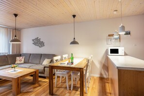 A modern living space with a wooden ceiling, a dining table, white chairs, a sofa, and a kitchen area with a microwave. - Apartment 'Ferienhof Paulsen - Regenpfeifer' with Shared Garden, Balcony and Wi-Fi (Pellworm)