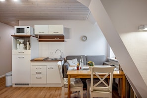 Une cuisine compacte avec des meubles blancs, un four à micro-ondes et un évier. On y trouve une table à manger en bois avec des chaises et un canapé gris. Une horloge est visible au mur.