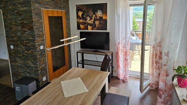Ein modernes Wohnzimmer mit Steinwand, Holztür, Flachbildfernseher, Esstisch und Blick durch eine Glastür.