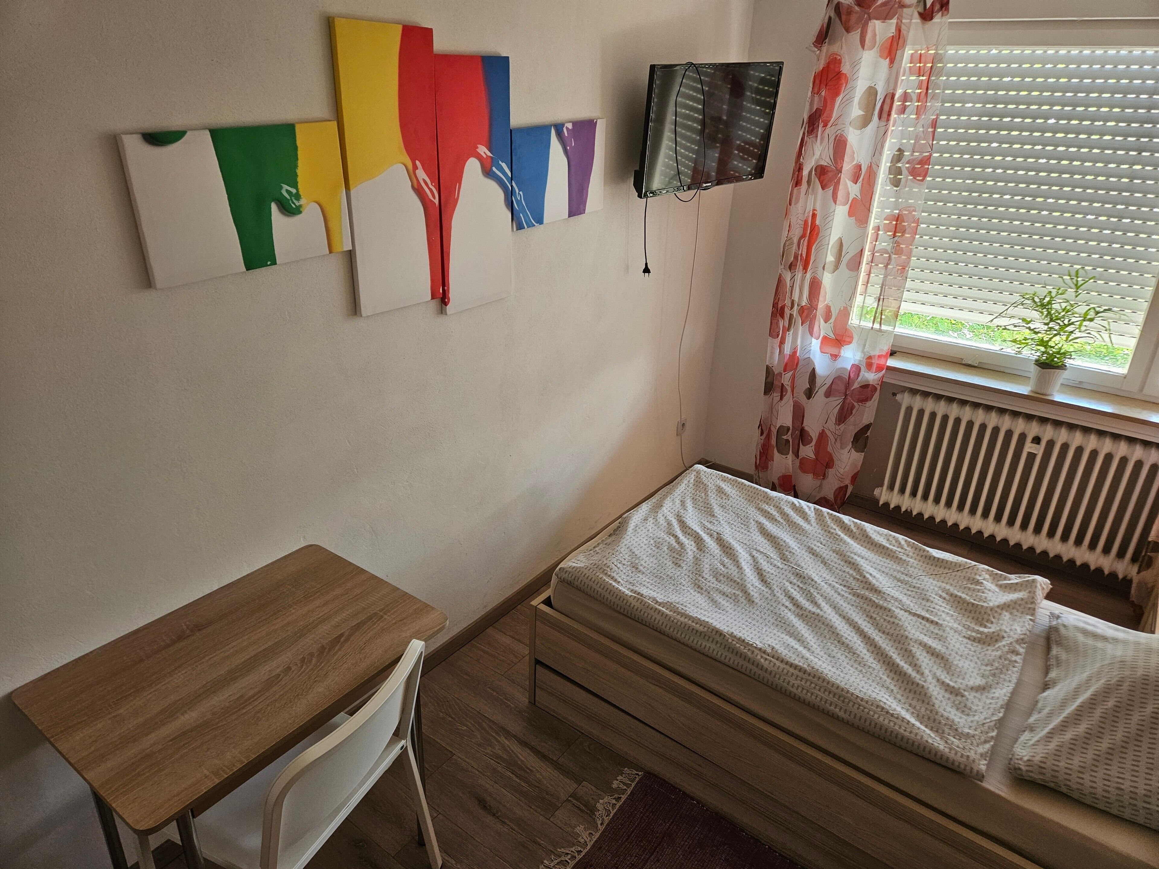 Una camera da letto con una scrivania in legno, un letto con biancheria bianca, un termosifone, una TV montata a parete e una finestra con tenda.