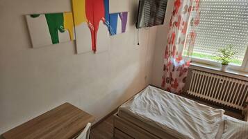 Una camera da letto con una scrivania in legno, un letto con biancheria bianca, un termosifone, una TV montata a parete e una finestra con tenda.
