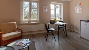Een kamer met een bank, een eettafel met stoelen, een bijzettafeltje met een tijdschrift, en een koelkast.