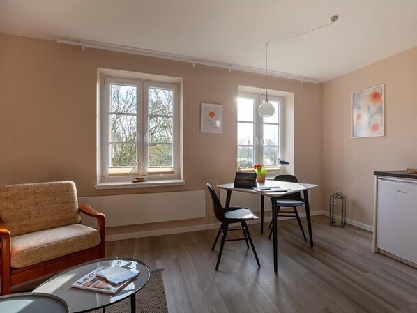 A room with a sofa, a dining table with chairs, a small table with a magazine, and a refrigerator.