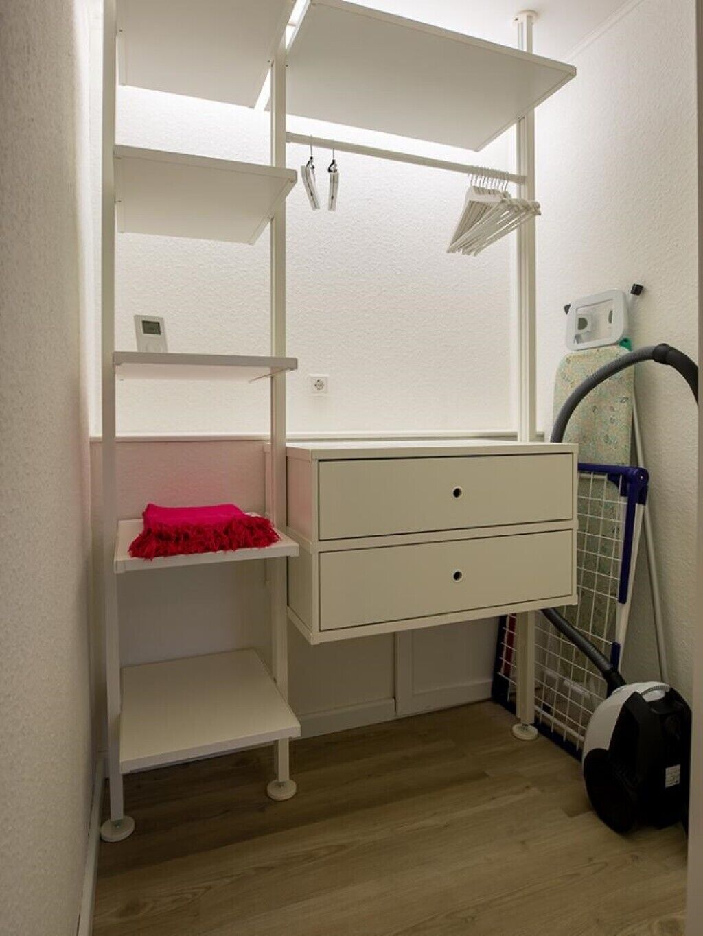 A compact room with a white wardrobe, a small red towel, and a vacuum cleaner.