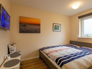 A bedroom with two beds, a TV, a painting of a sunset, a small table with a pillow, and a window.