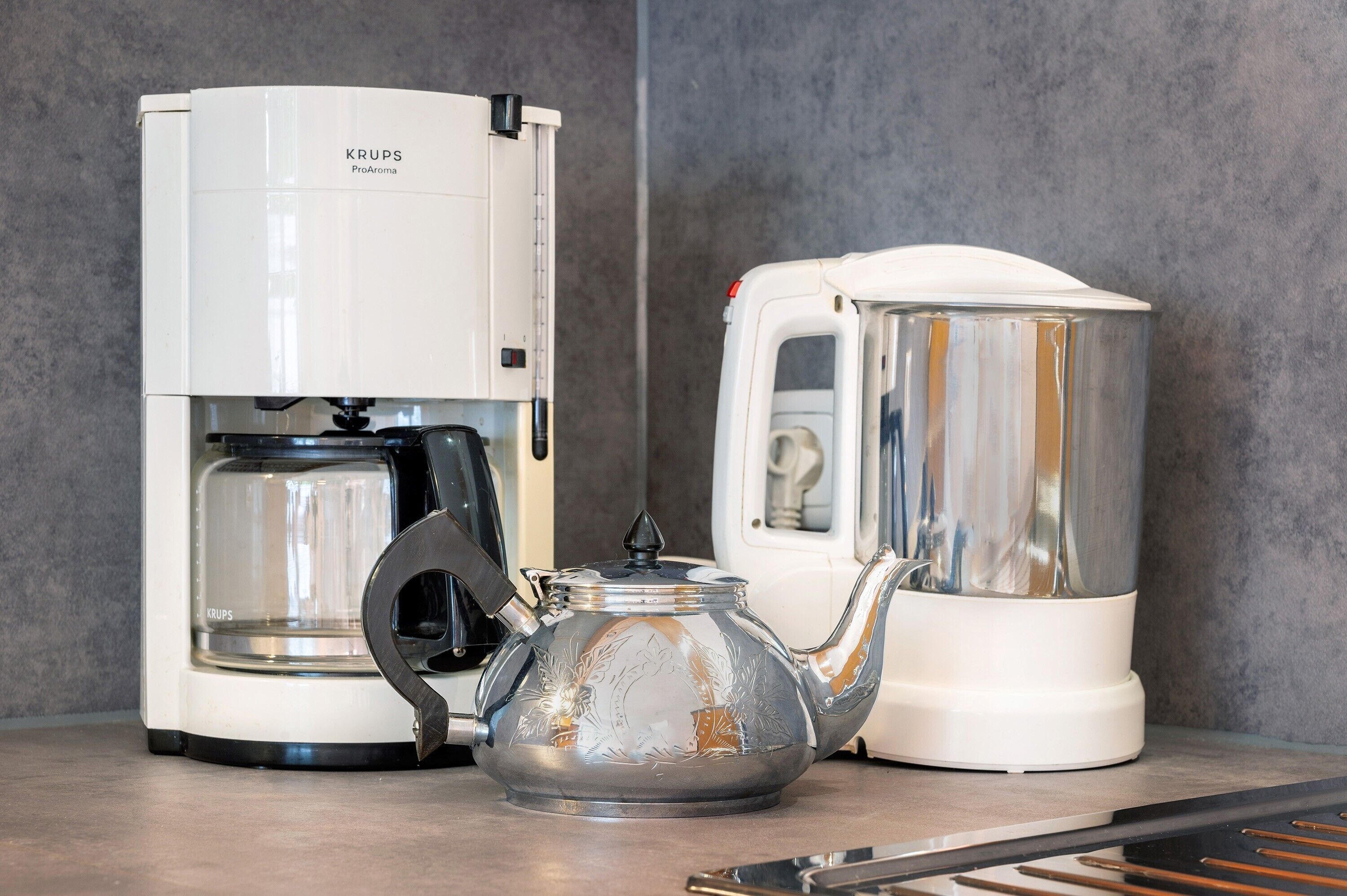 Eine Kaffeemaschine, eine Glas-Kaffeekanne und ein weißes Gerät auf einer Arbeitsplatte.