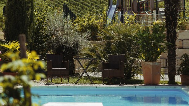 Un espace piscine avec un toboggan, entouré de verdure et bordé d’une colline où s’étendent des vignobles.