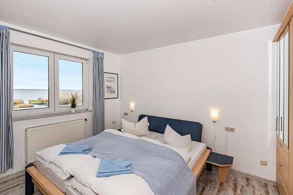 Ein Schlafzimmer mit Bett, Nachttischen, einem Fenster mit Vorhängen, einem Heizkörper und Meerblick.