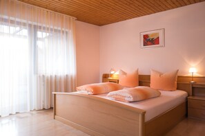 Un dormitorio con una cama de madera, dos mesitas de noche con lámparas, una ventana con cortinas translúcidas y un cuadro en la pared.