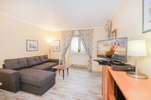 Un salon avec un canapé gris, une table basse en bois, un téléviseur posé sur un meuble et une fenêtre avec des rideaux.