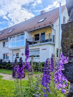 Un edificio de dos plantas con un balcón, un jardín con flores moradas y un cielo despejado.