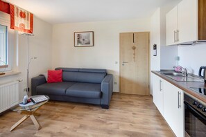 A compact living space with a grey sofa, a glass coffee table, a kitchenette with a stove, and a wooden door.