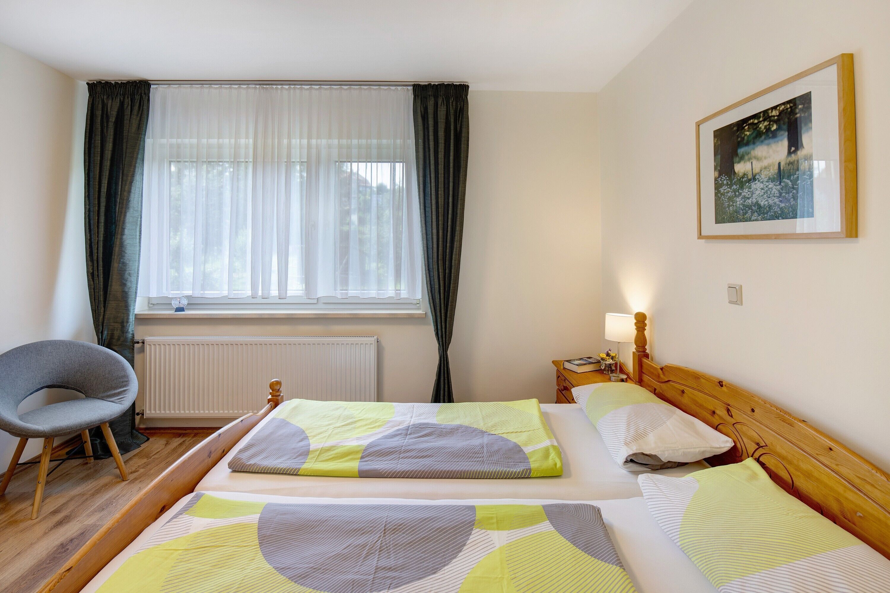 Camera d'albergo con due letti, una sedia, una finestra con tende, un termosifone, un comodino con una lampada e un quadro appeso al muro.