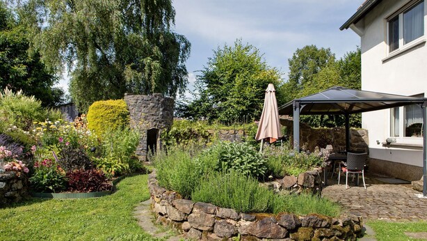Un jardín con un muro de piedra, un patio con mesa y sillas, y una zona de estar al aire libre techada.
